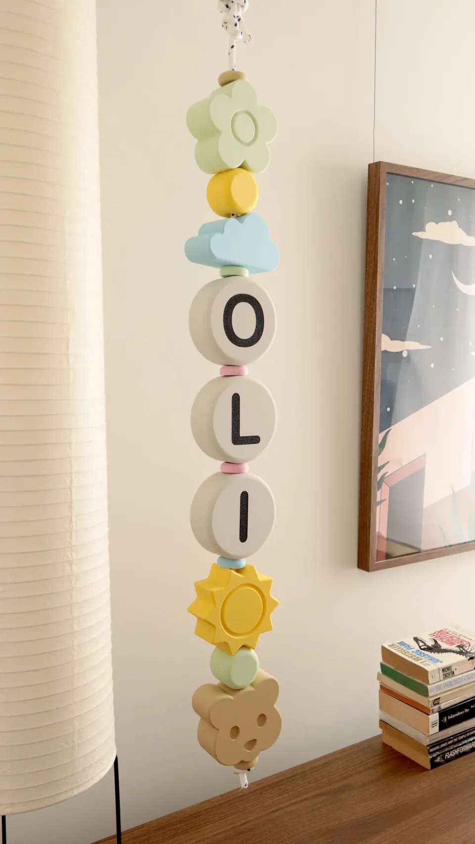 Guirnalda con el nombre OLI colgada en vertical en la pared de un cuarto, con flor, nube, sol y osito